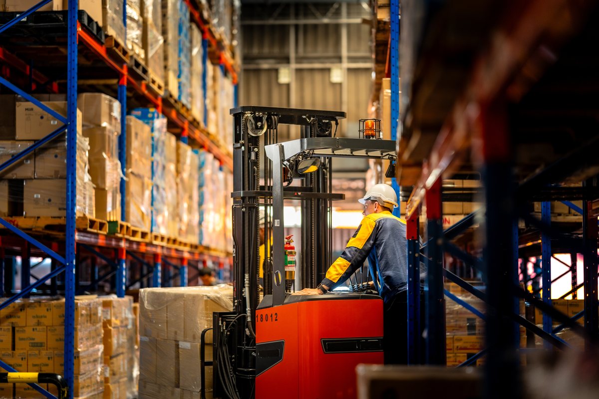 Operator on a red stand-up reach truck lifting a wrapped pallet in a narrow warehouse aisle between tall racking