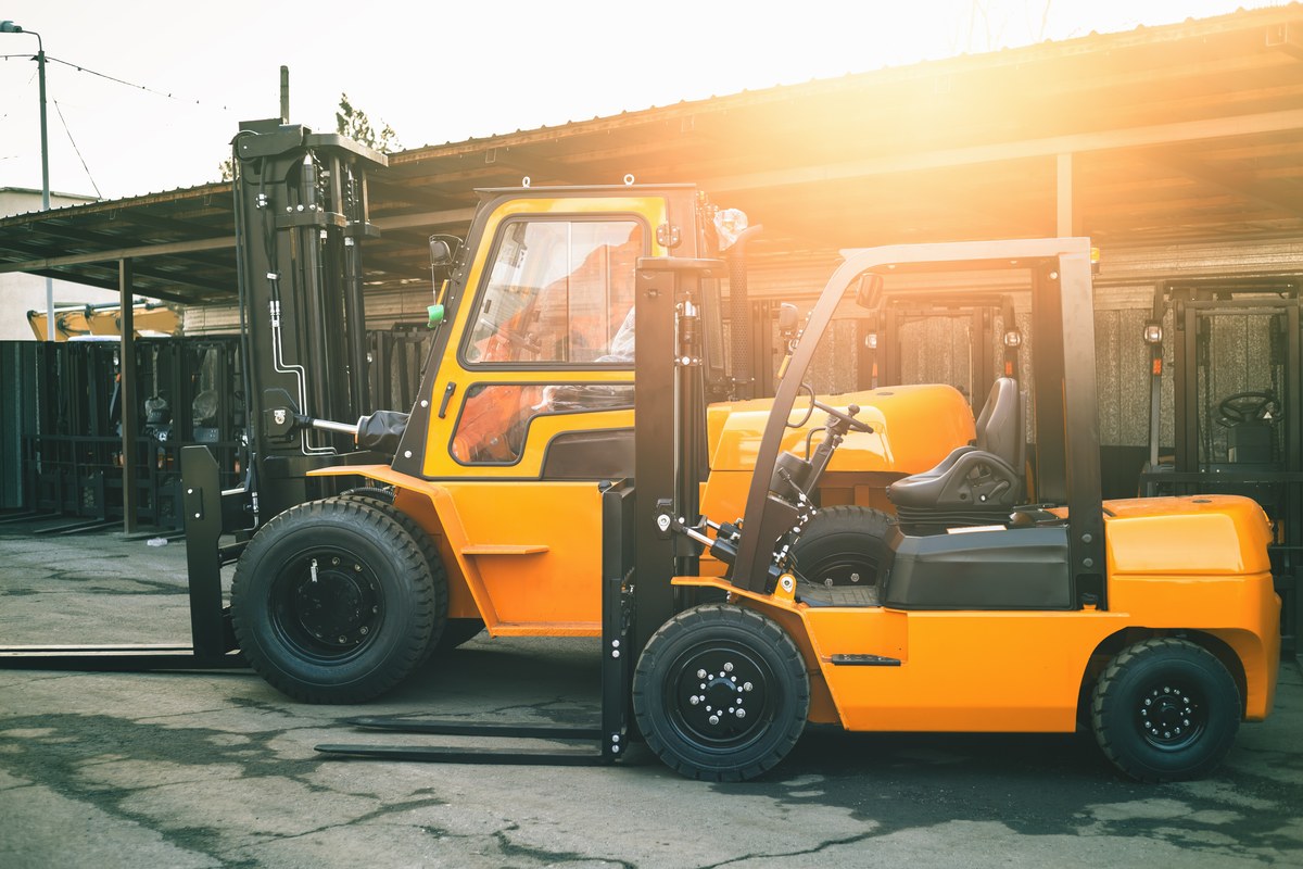Two new yellow sit-down forklifts parked on cracked pavement in a sunlit yard, the rear one with a cab and dual front tires, all on lug-tread tires mounted on black steel wheels