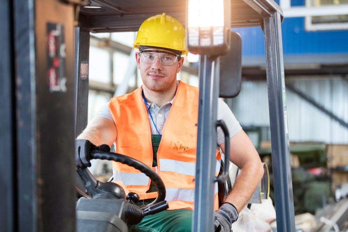 Forklift operator wearing an orange hi-vis vest, safety glasses and a yellow hard hat seated at the wheel of a lift truck