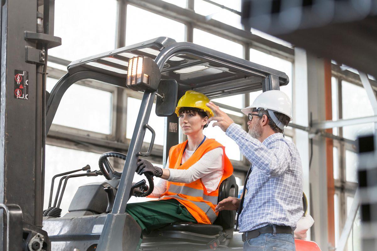 Supervisor in a white hard hat giving instructions to a forklift operator in an orange hi-vis vest and yellow hard hat seated at the controls of a lift truck