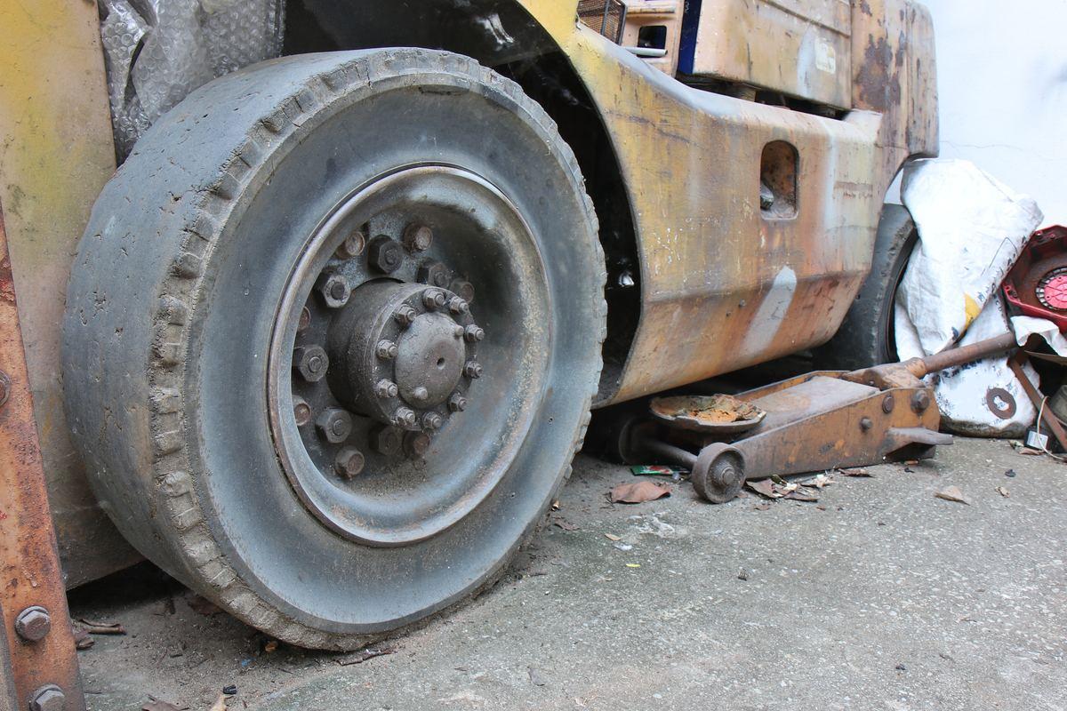 Front tire on an old counterbalance forklift worn smooth across the middle of the tread, with tread lugs left at the edges and a floor jack beside the truck