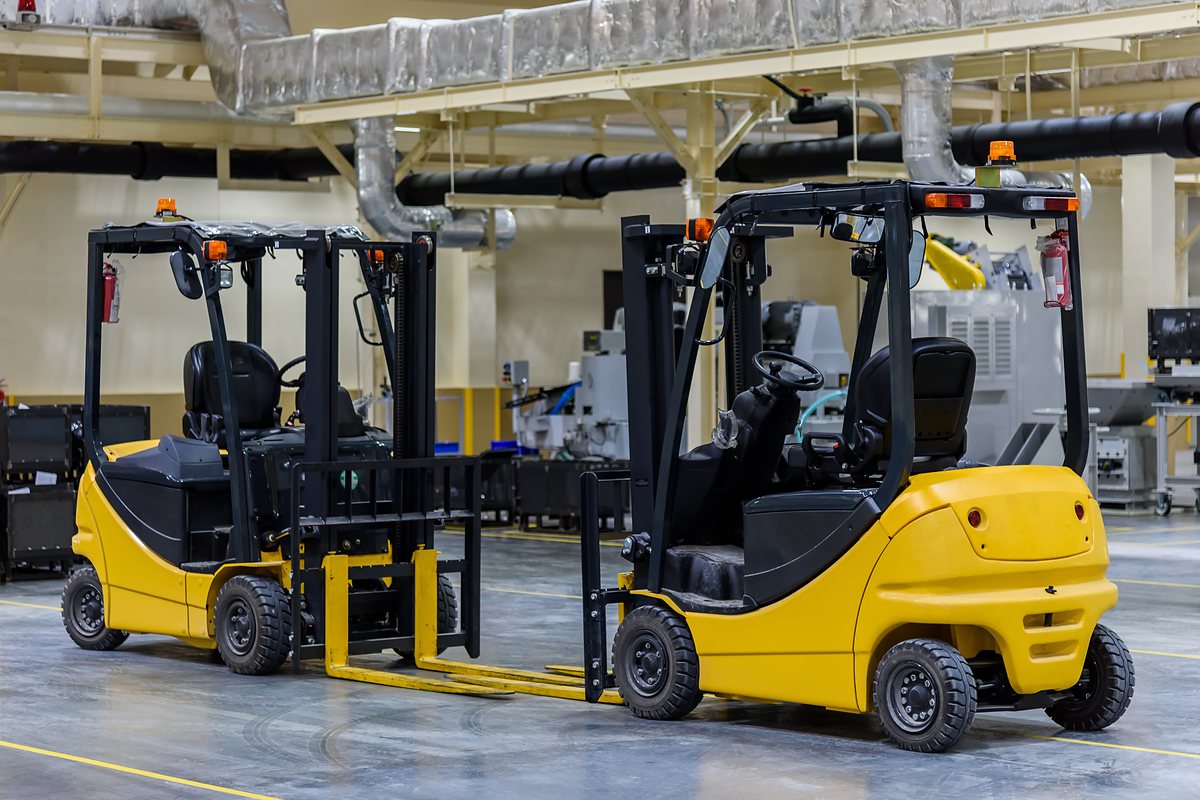 Two yellow forklifts parked on a factory floor beside industrial machines, with insulated ducts overhead